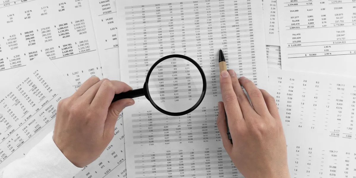 Woman holding a magnifying glass pointing at numbers on financial documents.