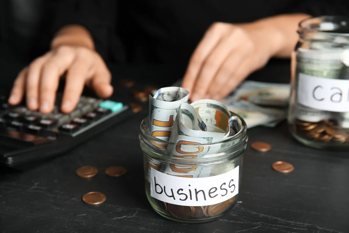 Woman using calculator near glass jar with money and tag BUSINESS on black table,